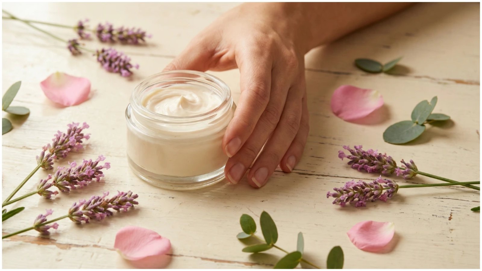 A glass jar of natural face cream surrounded by fresh lavender, rose petals, and eucalyptus leaves on a wooden table, representing gentle, plant-powered skincare in a non-toxic beauty routine.