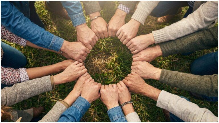 Circle of people standing outdoors with their fists touching in the center, forming a ring over grass to symbolize unity and mutual support in activism.